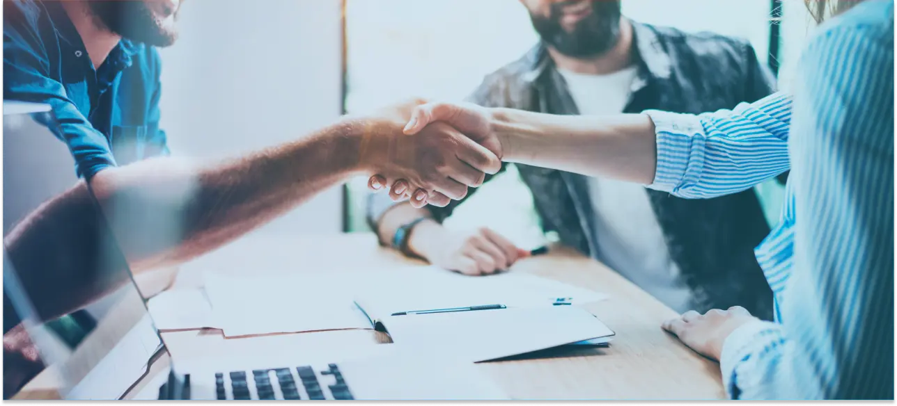 Two people shaking hands over a desk with documents and a laptop, symbolizing agreement or partnership, with others in the background.